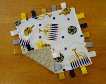 May include: A white baby blanket with a blue and gold menorah and star of David pattern. The blanket has multiple colorful ribbons attached to the edges.
