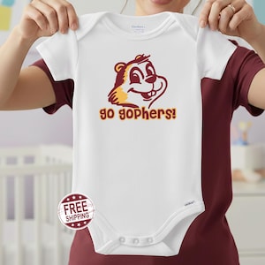 May include: White baby onesie with short sleeves featuring a cartoon gopher mascot in maroon and gold, with the text "go gophers!" in matching colors. The onesie is held up by a person.