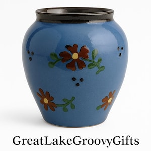 Puede incluir: Un pequeño jarrón de cerámica azul con un borde negro y diseños florales en tonos marrones, verdes y amarillos. El jarrón tiene un cuerpo redondo y una abertura ligeramente acampanada.