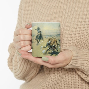 Może przedstawiać: Biały ceramiczny kubek z ilustracją w stylu vintage przedstawiającą kowboja na koniu łapiącego byka. Grafika przedstawia dynamiczną scenę z kowbojem, bykiem i psem w pustynnym krajobrazie. Kubek jest trzymany przez osobę.