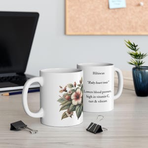 May include: Two white ceramic mugs. One mug features a floral design with hibiscus flowers in shades of pink, red, and green. The other mug has text that reads "Hibiscus, Ruby heart tonic, Lowers blood pressure, high in vitamin C, tart & vibrant."