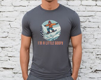 Puede incluir: Una camiseta gris oscuro con un gráfico de un snowboarder en una escena de invierno. El texto "I'm a little goofy" está impreso debajo del gráfico.