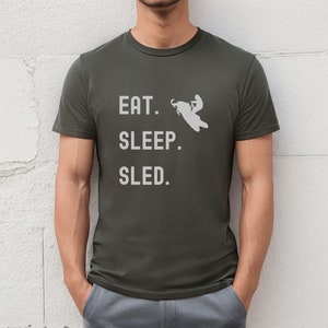 Puede incluir: Una camiseta verde oscuro con el texto "EAT. SLEEP. SLED." en blanco. Una silueta blanca de una persona conduciendo una moto de nieve est&aacute; por encima del texto.