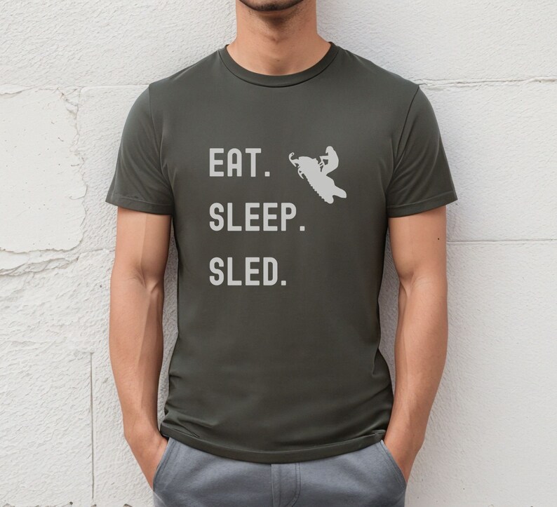 Puede incluir: Una camiseta verde oscuro con el texto "EAT. SLEEP. SLED." en blanco. Una silueta blanca de una persona conduciendo una moto de nieve est&aacute; por encima del texto.