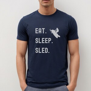 Puede incluir: Una camiseta azul marino con el texto "EAT. SLEEP. SLED." en letras blancas. Una silueta de una persona conduciendo una moto de nieve est&aacute; por encima del texto.