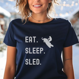 Puede incluir: Una camiseta azul marino con un gr&aacute;fico blanco de una persona conduciendo una moto de nieve. El texto "EAT. SLEEP. SLED." est&aacute; impreso en la camiseta.