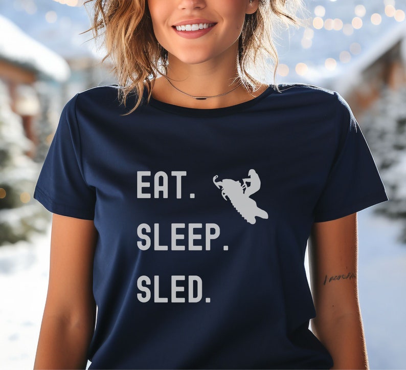 Puede incluir: Una camiseta azul marino con un gr&aacute;fico blanco de una persona conduciendo una moto de nieve. El texto "EAT. SLEEP. SLED." est&aacute; impreso en la camiseta.