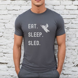 Puede incluir: Una camiseta gris oscuro con un gr&aacute;fico blanco de una persona conduciendo una moto de nieve. El texto "EAT. SLEEP. SLED." est&aacute; impreso en la camiseta.