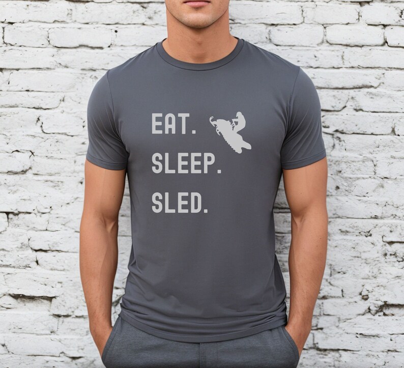 Puede incluir: Una camiseta gris oscuro con un gr&aacute;fico blanco de una persona conduciendo una moto de nieve. El texto "EAT. SLEEP. SLED." est&aacute; impreso en la camiseta.