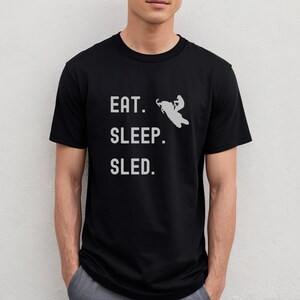 Puede incluir: Camiseta negra con el texto "EAT. SLEEP. SLED." en blanco. Una silueta de una persona conduciendo una moto de nieve est&aacute; por encima del texto.