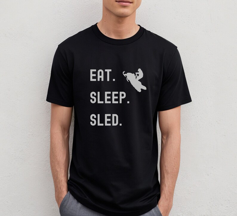 Puede incluir: Camiseta negra con el texto "EAT. SLEEP. SLED." en blanco. Una silueta de una persona conduciendo una moto de nieve est&aacute; por encima del texto.