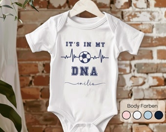 Może przedstawiać: Biały śpioszek dla niemowlęcia z niebieskim nadrukiem, na którym widnieje napis "It's in my DNA" wraz z piłką nożną i grafiką bicia serca. Imię "emilia" jest napisane kursywą pod grafiką. Śpioszek wisi na drewnianym wieszaku. Dostępne są cztery kolory śpioszka: różowy, biały, niebieski i czarny.