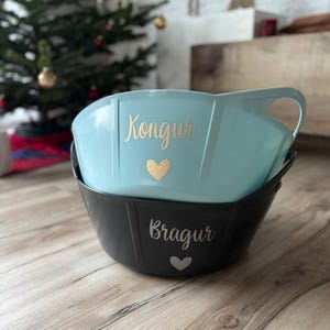 Könnte beinhalten: Zwei Plastik-Aufbewahrungsbehälter mit Griffen. Der obere Behälter ist hellblau mit dem Wort "Kongur" und einem goldenen Herzen. Der untere Behälter ist schwarz mit dem Wort "Bragur" und einem silbernen Herzen.