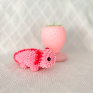 Puede incluir: Un juguete de peluche axolotl de crochet rosa y rojo con ojos negros. El axolotl est&aacute; sentado sobre una superficie blanca.