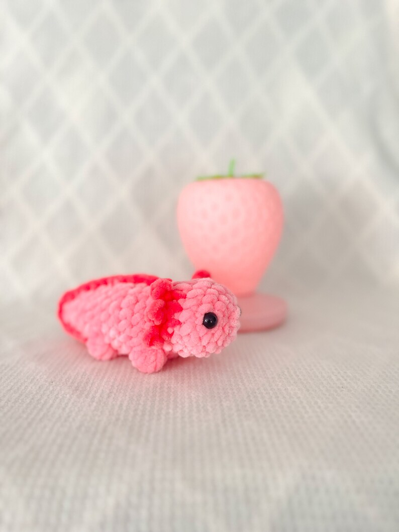Puede incluir: Un juguete de peluche axolotl de crochet rosa y rojo con ojos negros. El axolotl est&aacute; sentado sobre una superficie blanca.