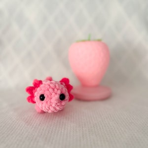 Puede incluir: Un juguete de peluche axolotl de crochet rosa con ojos negros. El juguete est&aacute; sentado sobre una superficie blanca.