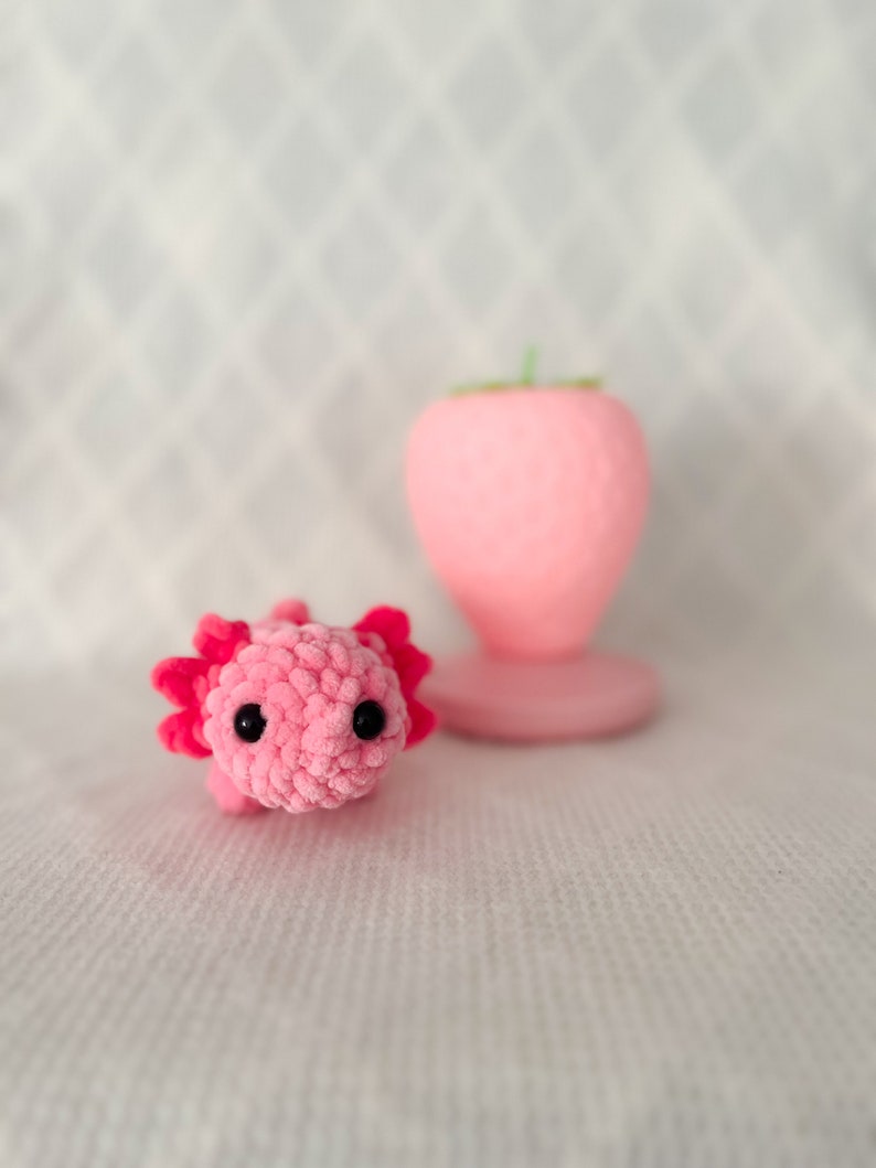 Puede incluir: Un juguete de peluche axolotl de crochet rosa con ojos negros. El juguete est&aacute; sentado sobre una superficie blanca.
