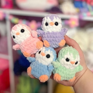 May include: Four crocheted penguin plushies in different colors: light pink, light blue, light green, and lavender. The penguins are all facing forward with their eyes closed and their beaks open.