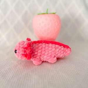 Puede incluir: Un juguete de peluche axolotl rosa con ojos negros. El axolotl est&aacute; acostado boca abajo con las patas extendidas. El juguete est&aacute; delante de un objeto rosa con forma de fresa.