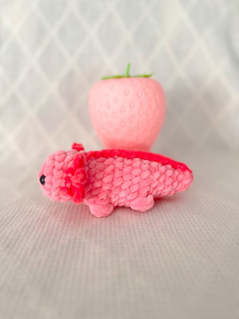 Puede incluir: Un juguete de peluche axolotl rosa con ojos negros. El axolotl est&aacute; acostado boca abajo con las patas extendidas. El juguete est&aacute; delante de un objeto rosa con forma de fresa.