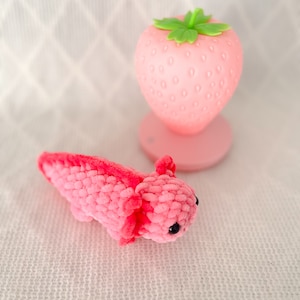 Puede incluir: Un juguete axolotl de crochet rosa con ojos negros. El axolotl est&aacute; acostado sobre una superficie blanca texturizada. Una luz nocturna en forma de fresa rosa con una hoja verde est&aacute; en el fondo.