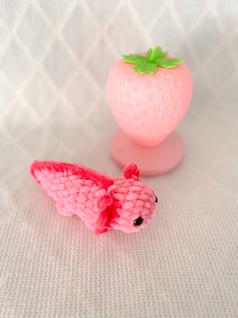 Puede incluir: Un juguete axolotl de crochet rosa con ojos negros. El axolotl est&aacute; acostado sobre una superficie blanca texturizada. Una luz nocturna en forma de fresa rosa con una hoja verde est&aacute; en el fondo.
