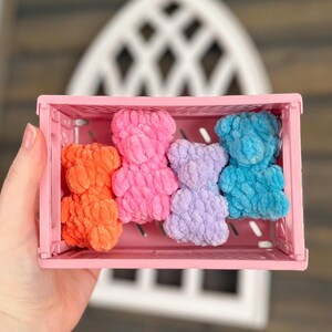 May include: Four crocheted teddy bears in a pink plastic crate. The bears are in shades of orange, pink, purple, and blue.