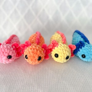 Puede incluir: Cuatro peluches de axolotl tejidos a crochet en rosa, naranja, amarillo y azul. Los axolotls est&aacute;n todos mirando a la c&aacute;mara y tienen ojos negros.
