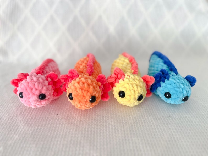 Puede incluir: Cuatro peluches de axolotl tejidos a crochet en rosa, naranja, amarillo y azul. Los axolotls est&aacute;n todos mirando a la c&aacute;mara y tienen ojos negros.