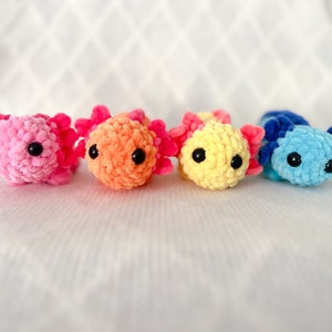 Puede incluir: Cuatro peluches de axolotl de crochet en diferentes colores: rosa, naranja, amarillo y azul. Cada axolotl tiene ojos negros y una textura esponjosa.