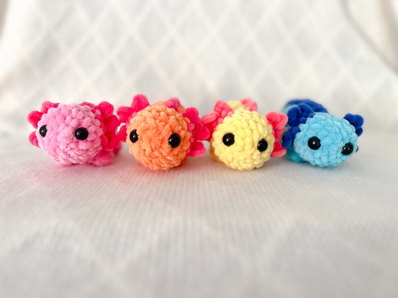 Puede incluir: Cuatro peluches de axolotl de crochet en diferentes colores: rosa, naranja, amarillo y azul. Cada axolotl tiene ojos negros y una textura esponjosa.