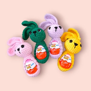 May include: Four crocheted bunny rabbits in pink, green, purple, and yellow holding Kinder Joy chocolate eggs. Each bunny has a different colored egg inside its body.
