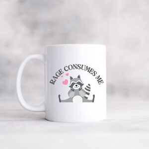 Peut inclure: Une tasse &agrave; caf&eacute; en c&eacute;ramique blanche avec un raton laveur de dessin anim&eacute; noir et gris portant un n&oelig;ud papillon rose. La tasse porte le texte "RAGE CONSUMES ME" en noir autour du raton laveur.