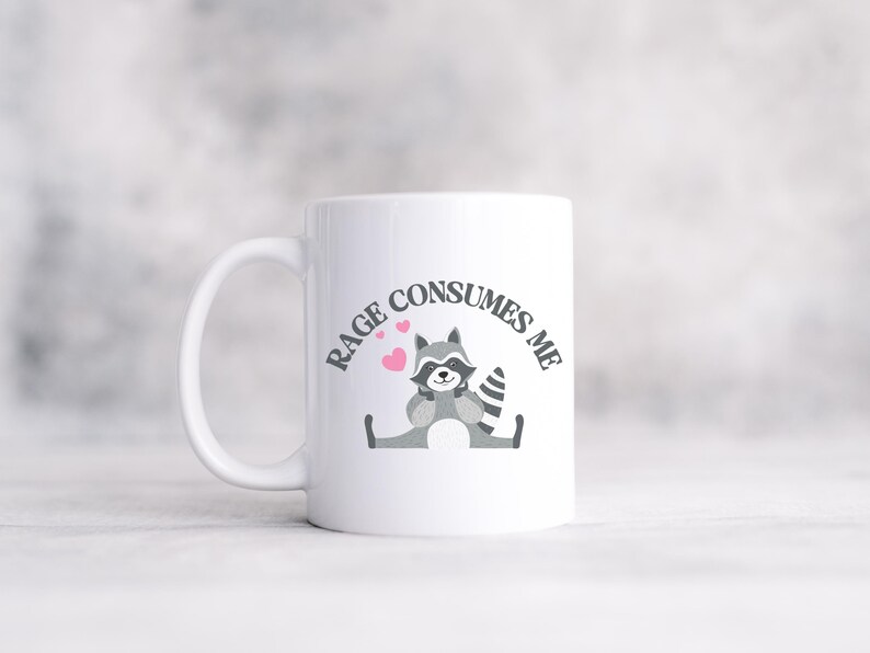 Peut inclure: Une tasse &agrave; caf&eacute; en c&eacute;ramique blanche avec un raton laveur de dessin anim&eacute; noir et gris portant un n&oelig;ud papillon rose. La tasse porte le texte "RAGE CONSUMES ME" en noir autour du raton laveur.
