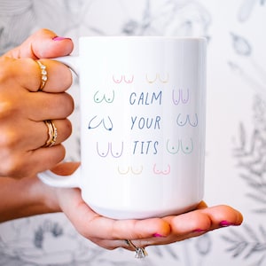 May include: White ceramic mug with the text "CALM YOUR TITS" in blue, green, and purple. The mug features colorful illustrations of breasts. The mug is held by a person with multiple rings on their fingers.