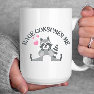 Peut inclure: Une tasse &agrave; caf&eacute; en c&eacute;ramique blanche avec une illustration de raton laveur noir et gris et le texte "Rage Consumes Me" en arc gris au-dessus du raton laveur. Le raton laveur tient ses pattes ensemble et a des c&oelig;urs roses autour de lui.