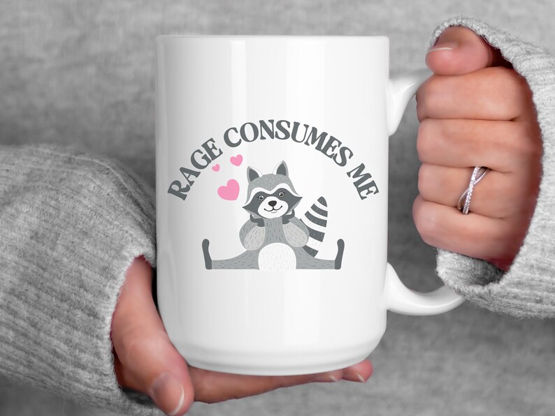 Peut inclure: Une tasse &agrave; caf&eacute; en c&eacute;ramique blanche avec une illustration de raton laveur noir et gris et le texte "Rage Consumes Me" en arc gris au-dessus du raton laveur. Le raton laveur tient ses pattes ensemble et a des c&oelig;urs roses autour de lui.
