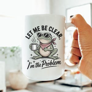 May include: A white ceramic mug featuring a graphic of a green frog wearing sunglasses and a pink neckerchief, holding a small cup. The text reads, "LET ME BE CLEAR, I'm the Problem," with small star accents surrounding the illustrated frog character.