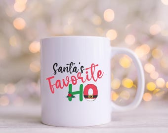 May include: White ceramic mug with the text "Santa's Favorite HO" in black and red script. The "HO" is designed with a Santa belt and holly. The mug has a white handle and is set against a blurred background of lights.