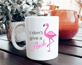 May include: White ceramic mug with a pink flamingo illustration and the phrase "I don't give a Flock" in black and pink script. The mug has a curved handle and is set on a dark wooden surface.