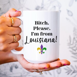 May include: White ceramic mug with the text "Bitch. Please. I'm from Louisiana!" in black script. The mug features a fleur-de-lis symbol in purple, gold, and green. The mug is held by a person with rings on their fingers.