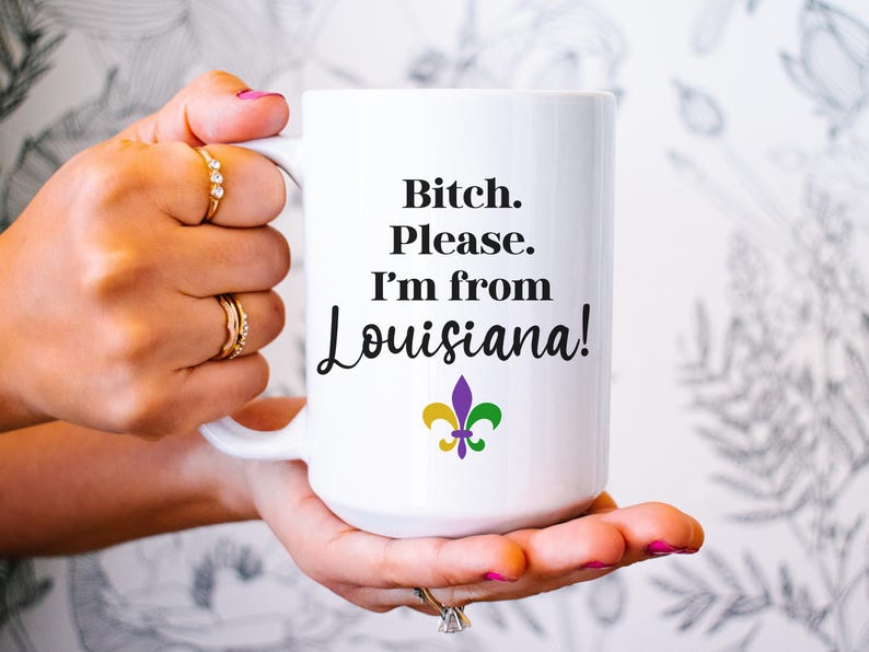 May include: White ceramic mug with the text "Bitch. Please. I'm from Louisiana!" in black script. The mug features a fleur-de-lis symbol in purple, gold, and green. The mug is held by a person with rings on their fingers.