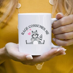 Peut inclure: Une tasse &agrave; caf&eacute; en c&eacute;ramique blanche avec une illustration de raton laveur noir et gris et le texte "Rage Consumes Me".