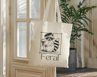 May include: A white canvas tote bag with black handles. The bag features a black and white illustration of a raccoon wearing a beret and holding a glass of wine. The text "Feral" is printed in black below the illustration.