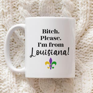 May include: White ceramic mug with the text "Bitch. Please. I'm from Louisiana!" in black script. A fleur-de-lis in purple, green, and yellow is below the text. The mug has a white handle.