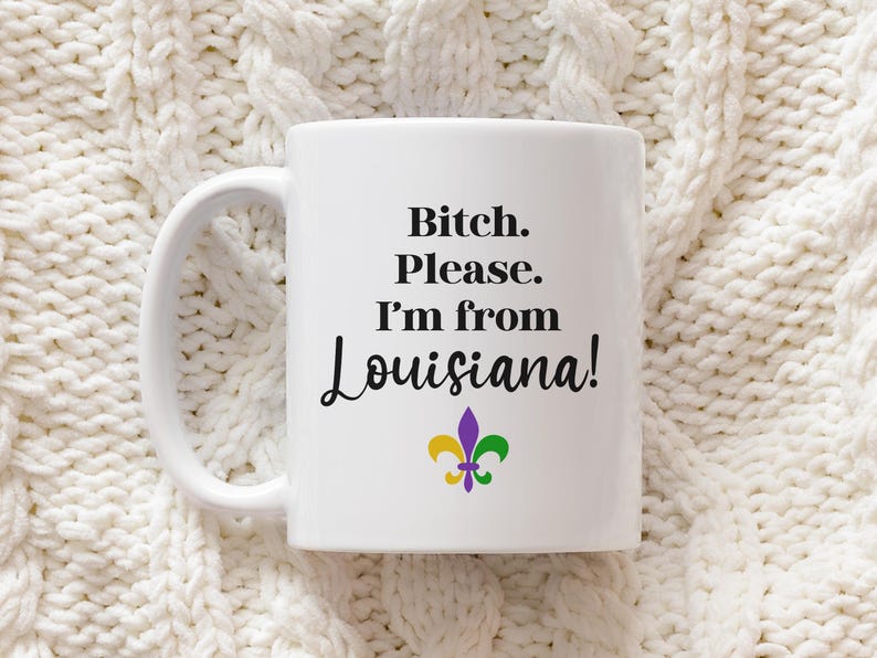 May include: White ceramic mug with the text "Bitch. Please. I'm from Louisiana!" in black script. A fleur-de-lis in purple, green, and yellow is below the text. The mug has a white handle.