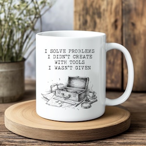 May include: A white ceramic mug featuring a black ink-style illustration of an open toolbox, a ruler, a magnifying glass, and a cup of coffee. The text printed on the mug reads: I SOLVE PROBLEMS I DIDN'T CREATE WITH TOOLS I WASN'T GIVEN.