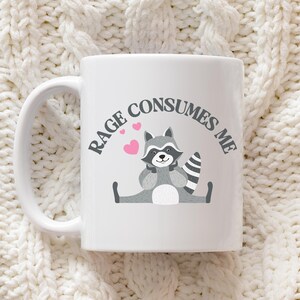 Peut inclure: Une tasse en c&eacute;ramique blanche avec une illustration de raton laveur noir et gris et le texte "Rage Consumes Me".