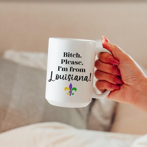 May include: White ceramic mug with the text "Bitch. Please. I'm from Louisiana!" in black script. A fleur-de-lis in purple, green, and yellow is below the text. The mug is held by a person with red nail polish.