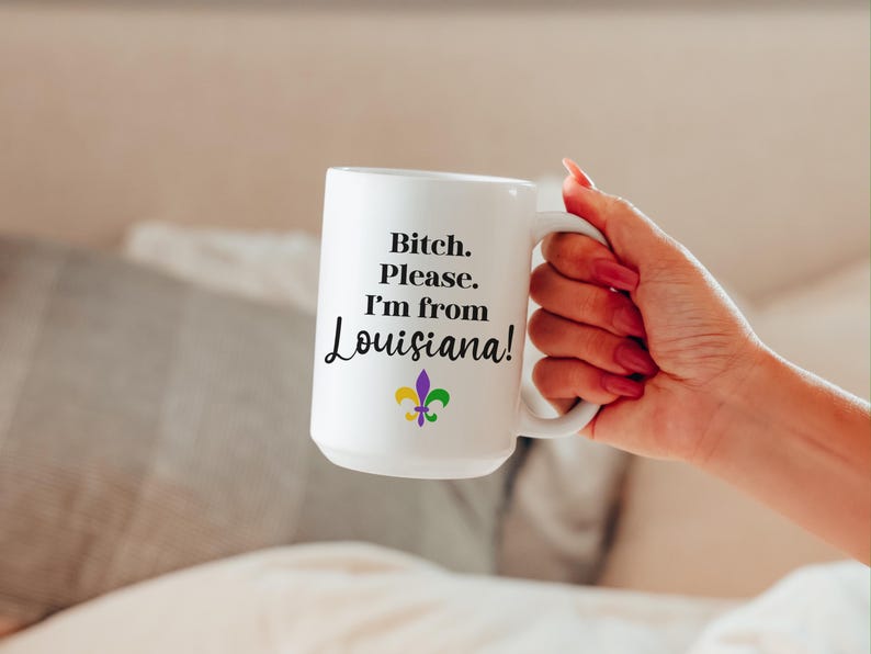 May include: White ceramic mug with the text "Bitch. Please. I'm from Louisiana!" in black script. A fleur-de-lis in purple, green, and yellow is below the text. The mug is held by a person with red nail polish.